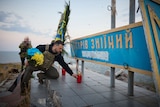 Zelenskyy carrying blue and yellow flowers kneeling over steps and placing red candle infront of sign
