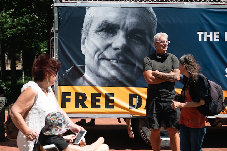 A man stands, arms folded, in front of a large banner with his face on it and the words 'Free David'. 
