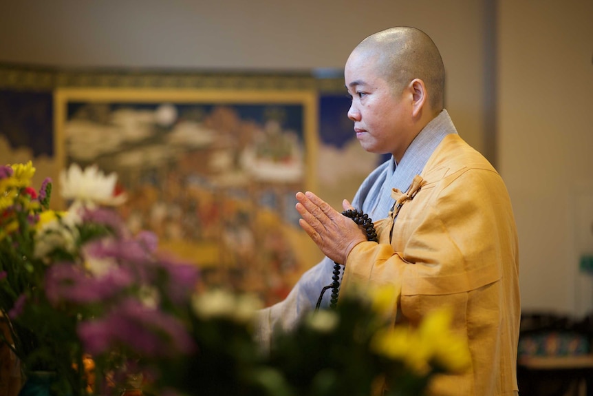 A buddhist nun in yellow and grey robes stands holding black beads at a memorial site adorned with multicolour flowers.