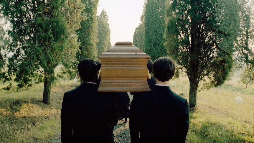 A group of men carry a wooden coffin along a tree-lined avenue.