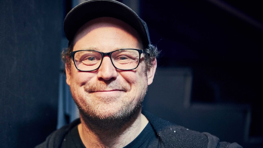 Ben Wellwood smiling, he is wearing glasses, a baseball cap and a black hoodie.