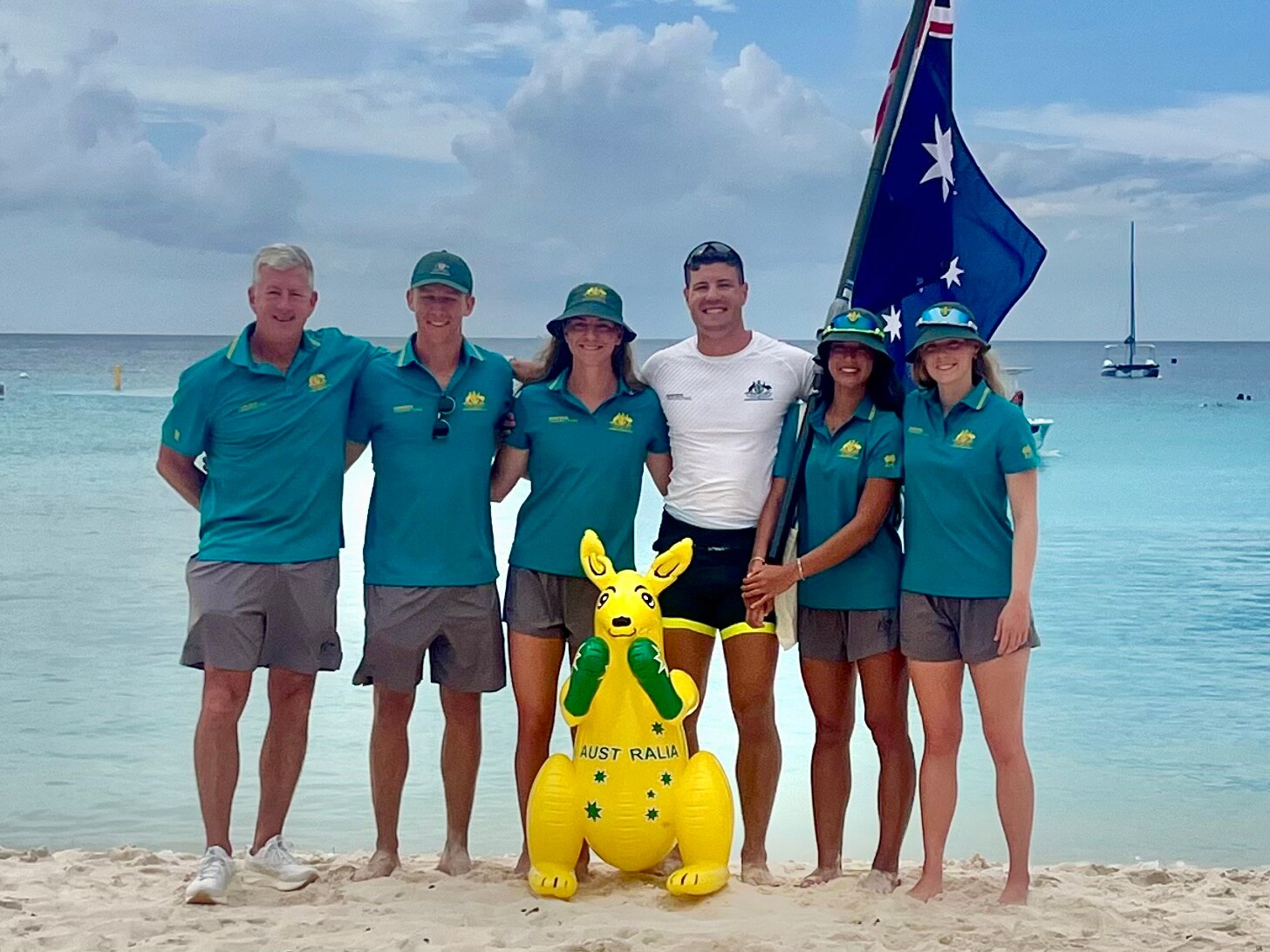 Cinco personas con uniformes deportivos australianos y un hombre con camisa blanca se encuentran en una playa con una bandera australiana 