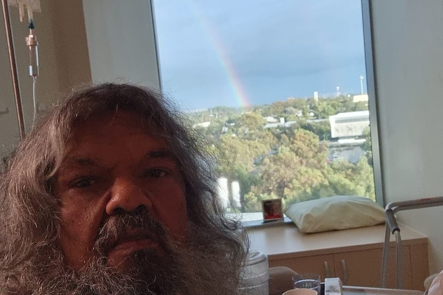 A man sits in bed and takes a selfie with a rainbow visible out the window
