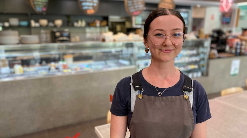 A young woman with an apron on smiles at the camera, with a cafe cake fridge behind