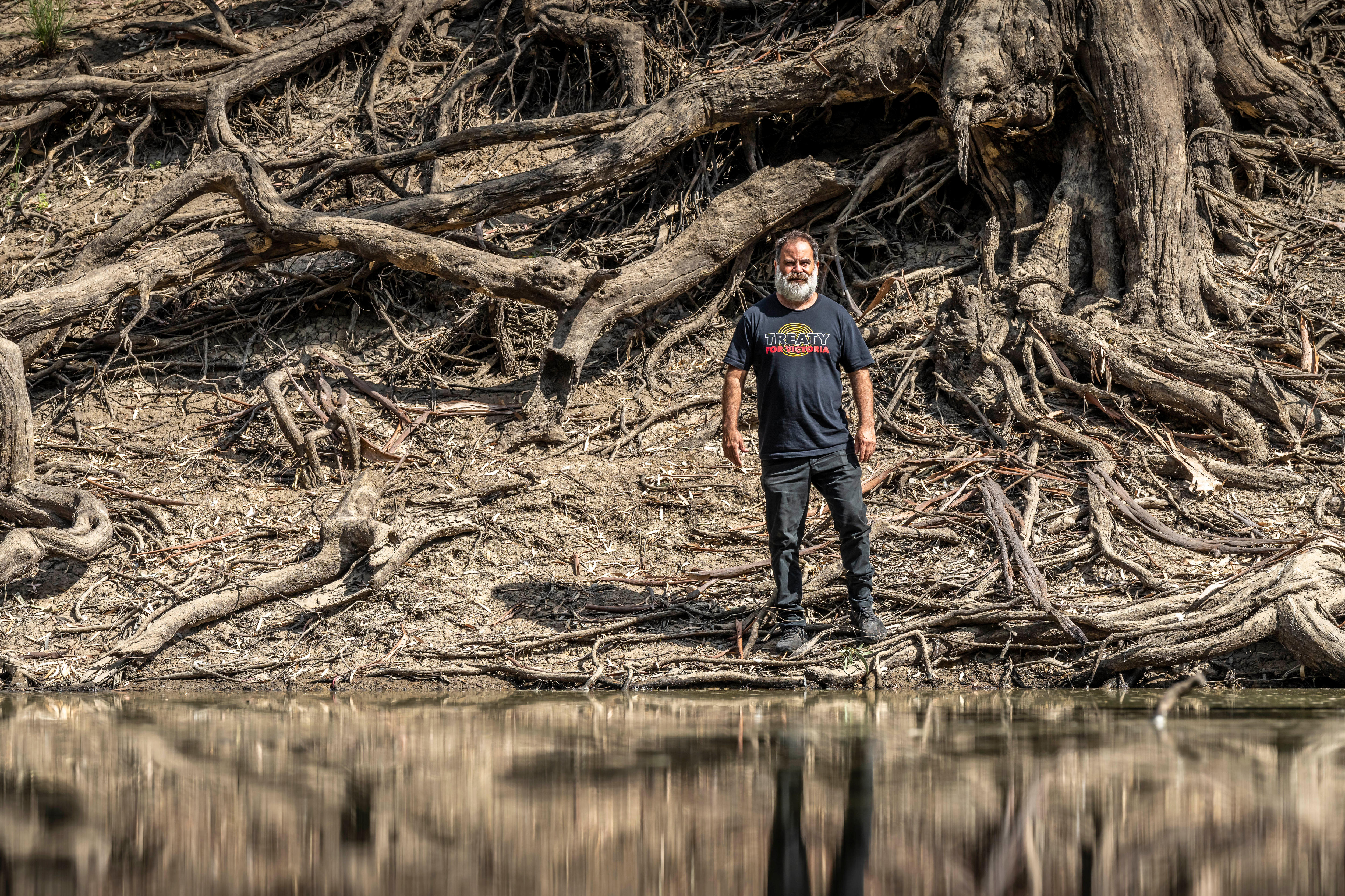 First Nations man wearing close the gap shirt stands in river, in front of huge tree roots on a riverbank