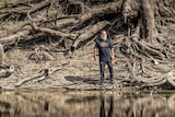 First Nations man wearing close the gap shirt stands in river, in front of huge tree roots on a riverbank