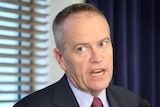 Bill Shorten, wearing a maroon tie and pale blue shirt, speaks at a lectern. There are venetian blinds on the window behind him