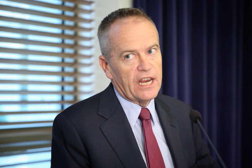 Bill Shorten, wearing a maroon tie and pale blue shirt, speaks at a lectern. There are venetian blinds on the window behind him