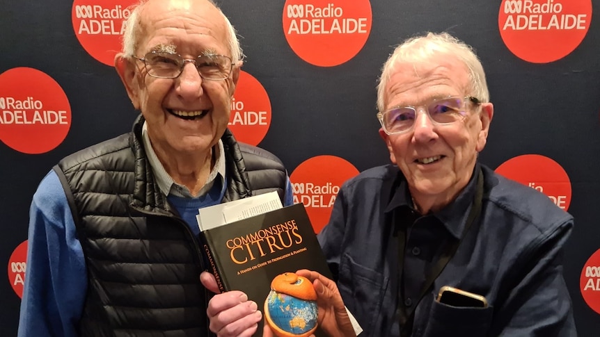 two older men with grey hair and glasses smiling infront of pull-up banner with ABC logo on it, holding book