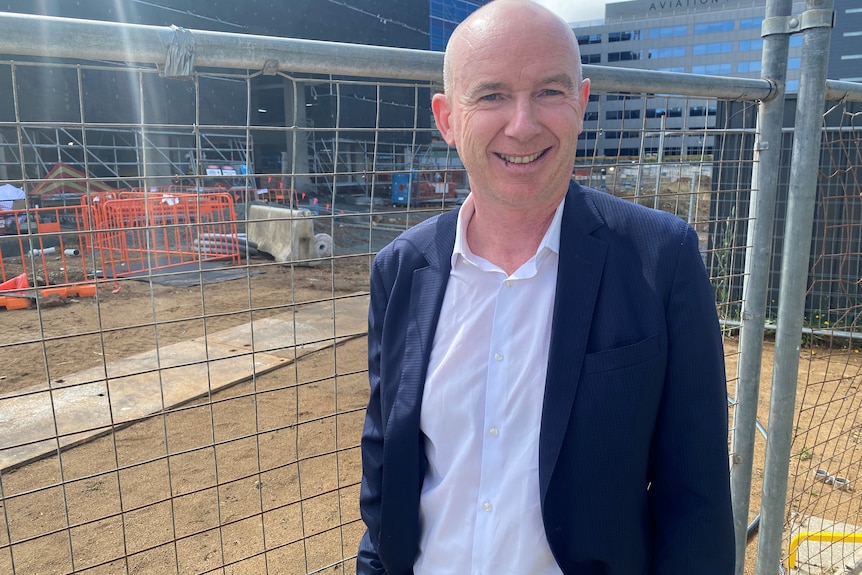 A man in a suit jacket smiles in front of a construction site.