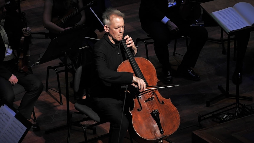 Cellist Alban Gerhardt playing on a darkened stage, with a spotlight on him