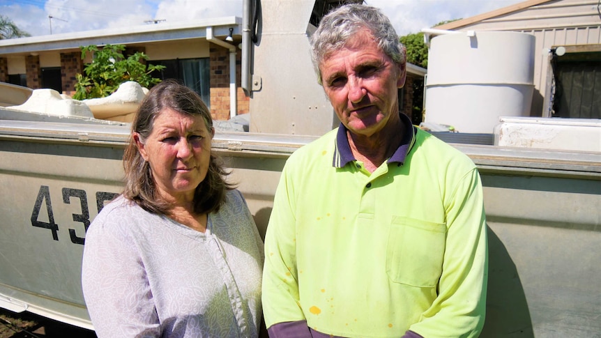 An older couple with greying hair stand solemn in front of their boat - their arms crossed. The man still in his work clothes