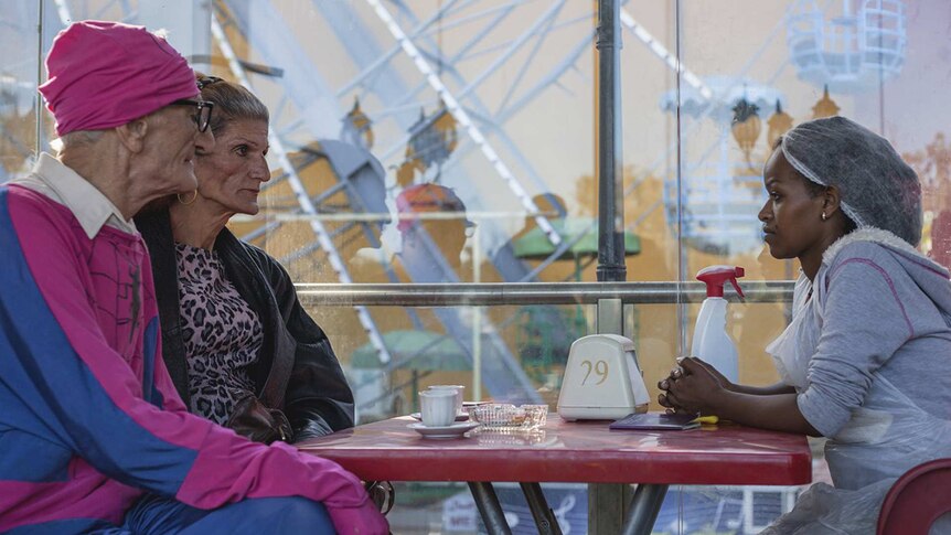 Colour photo of Yordanos Shiferaw sitting at a table opposite two people in an amusement park in 2018 film Capharnaüm.
