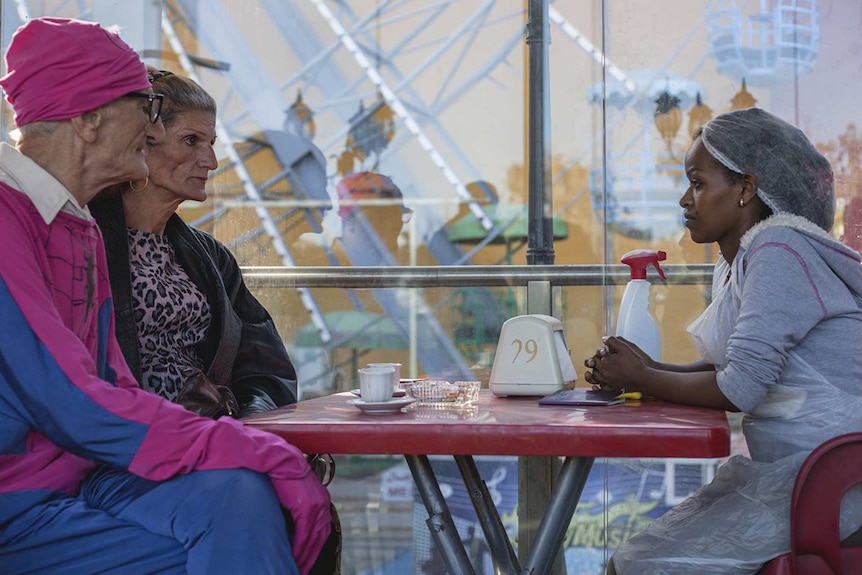 Colour photo of Yordanos Shiferaw sitting at a table opposite two people in an amusement park in 2018 film Capharnaüm.