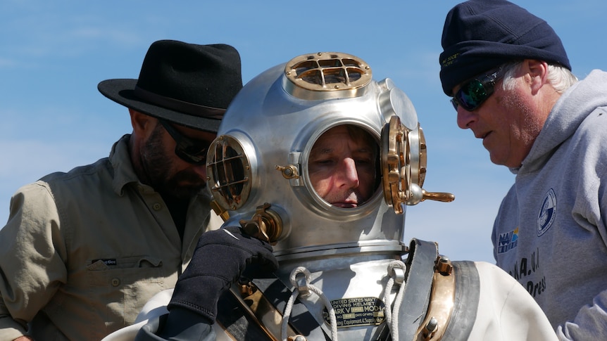 A close-up of a copper diving helmet with the window open as two men help tighten screws on the chest plate.