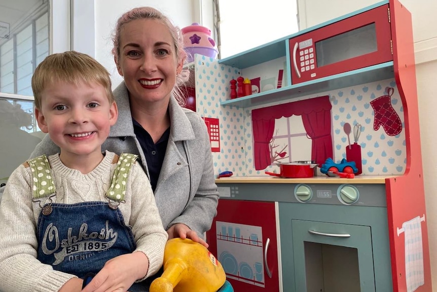 woman with boy sitting on lap play toy kitchen in background