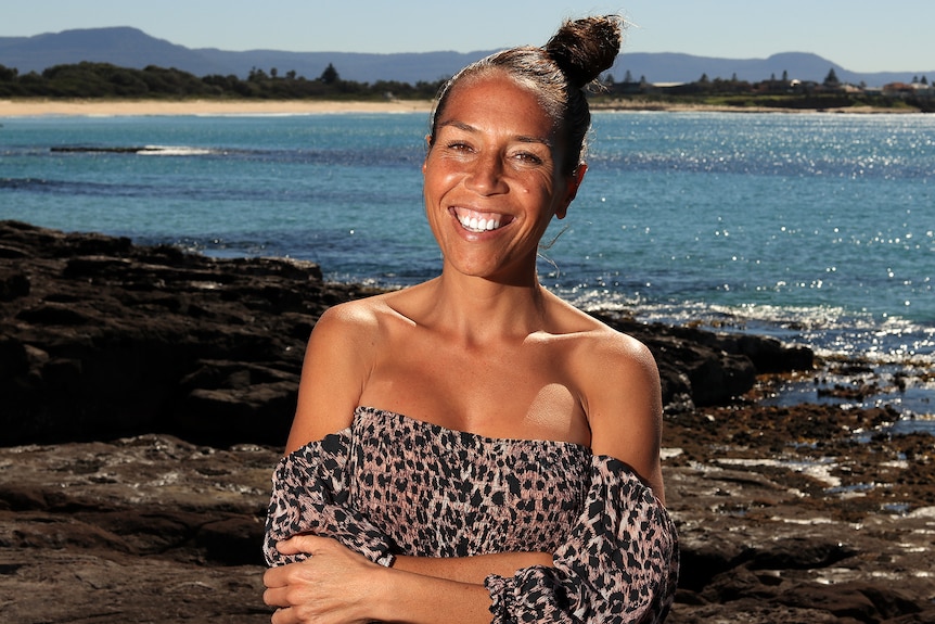 A young woman wearing a dress and smiling on a rock shelf in front of sparkling blue water.