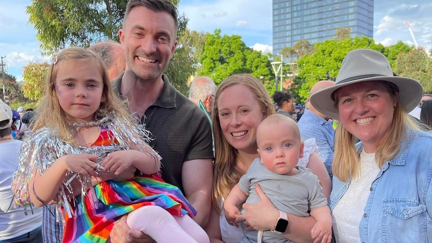 man holding young girl in rainbow dress and two women with baby boy in grey t-shirt smiling