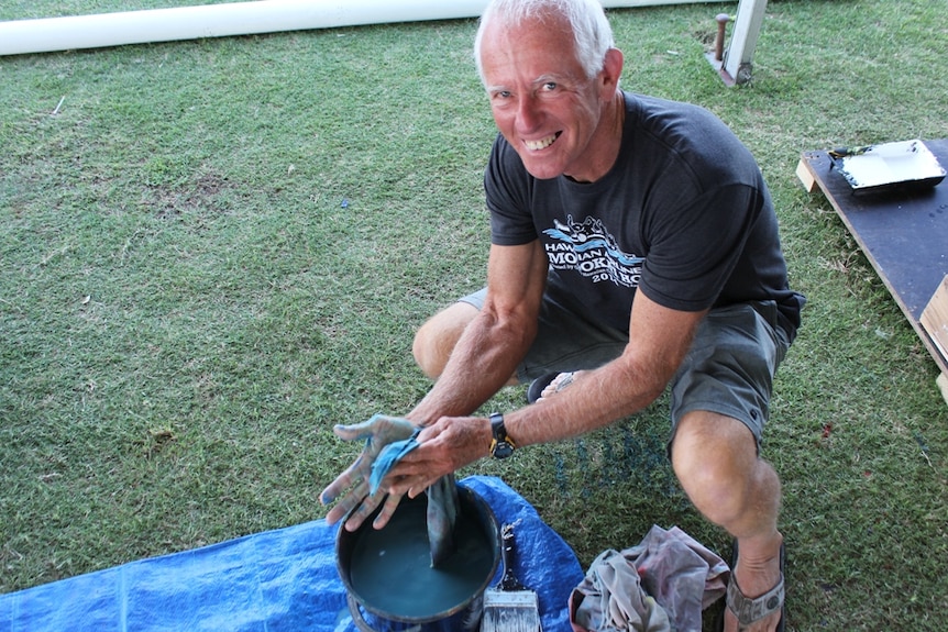 An older man is smiling, kneeling at bucket of water where he washes paint from his hands.