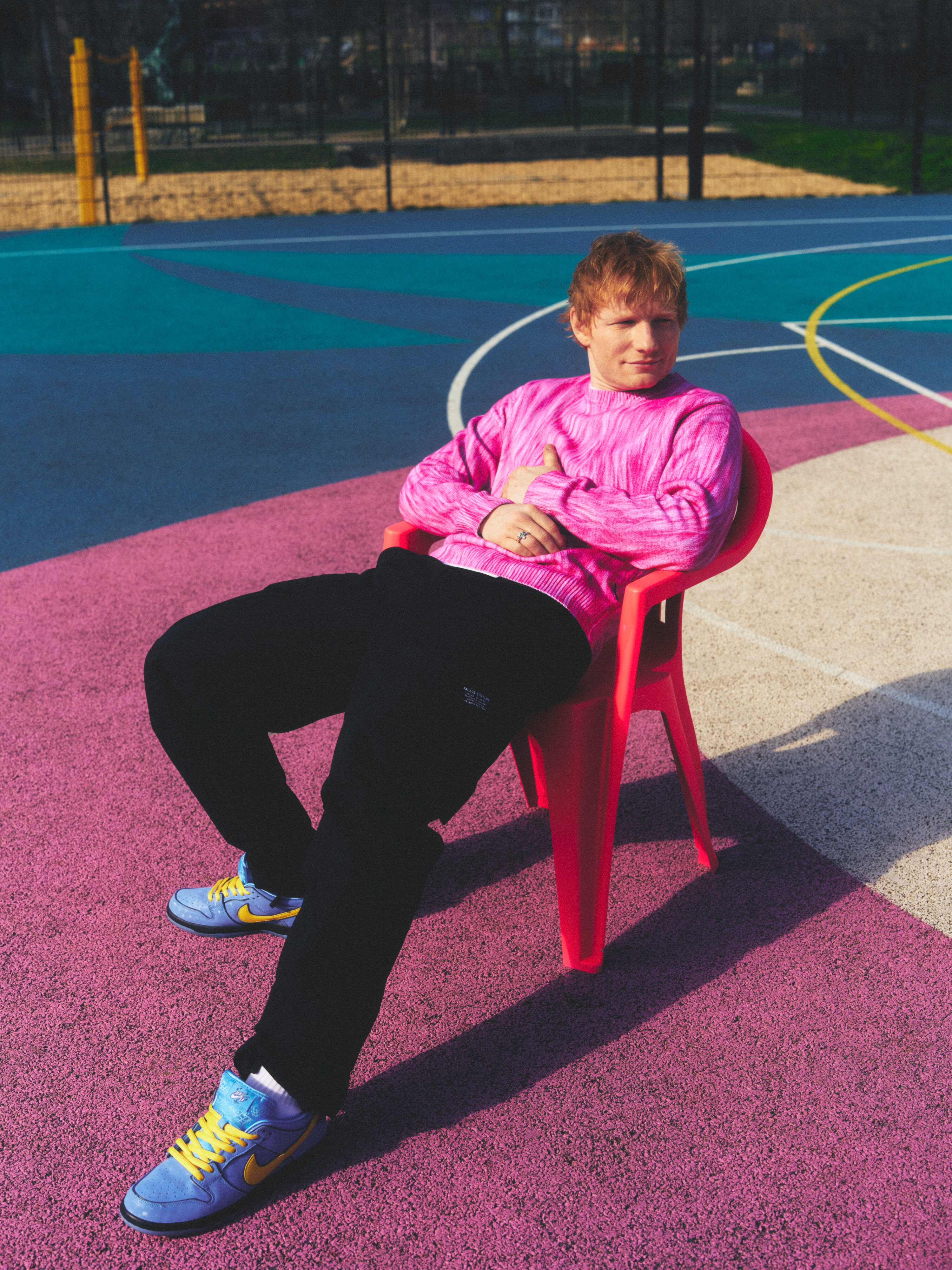 Ed Sheeran sits on a pink plastic chair in a pink jumper. 