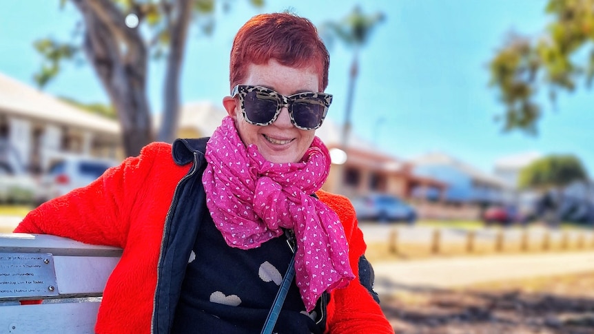 Linda smiling wearing a red jacket, bright pink scarf, big sunglasses and short hair after her surgery.