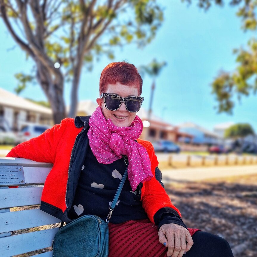 Linda smiling wearing a red jacket, bright pink scarf, big sunglasses and short hair after her surgery.