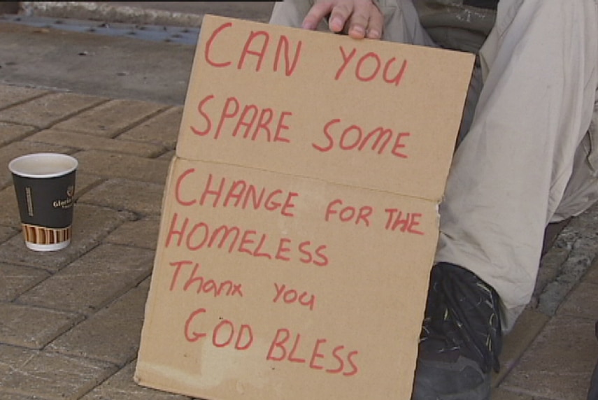 A homeless person, face not visible, sits on a street holding a cardboard sign asking for money.