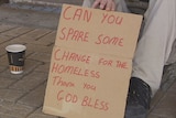 A homeless person, face not visible, sits on a street holding a cardboard sign asking for money.