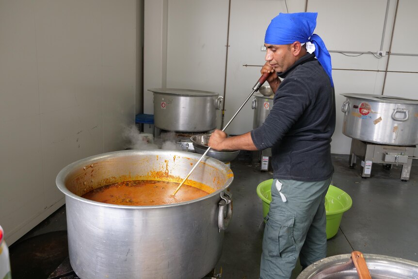 A man wearing a blue head scarf stirring a large pot of food with a stick