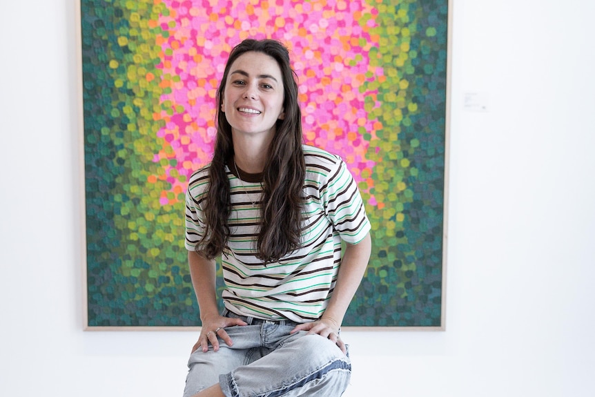Une jeune femme est assise devant un tableau coloré.