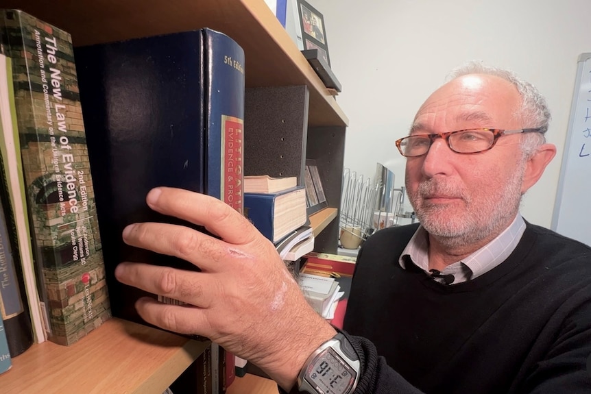 A man with glasses places a thick legal book on a shelf in an office. He has a neutral expression on his face.