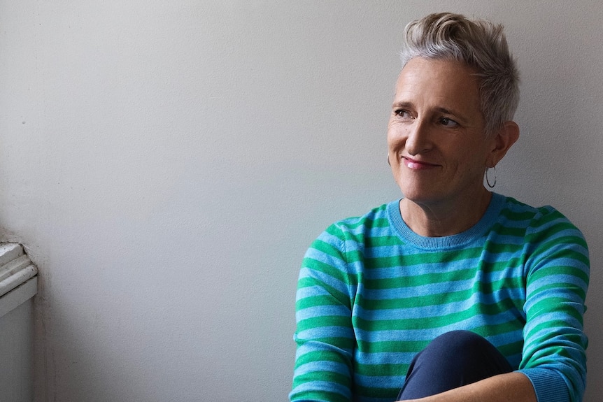 A woman with short grey hair and a blue and green striped top sits with her hands on her knees looking to the side