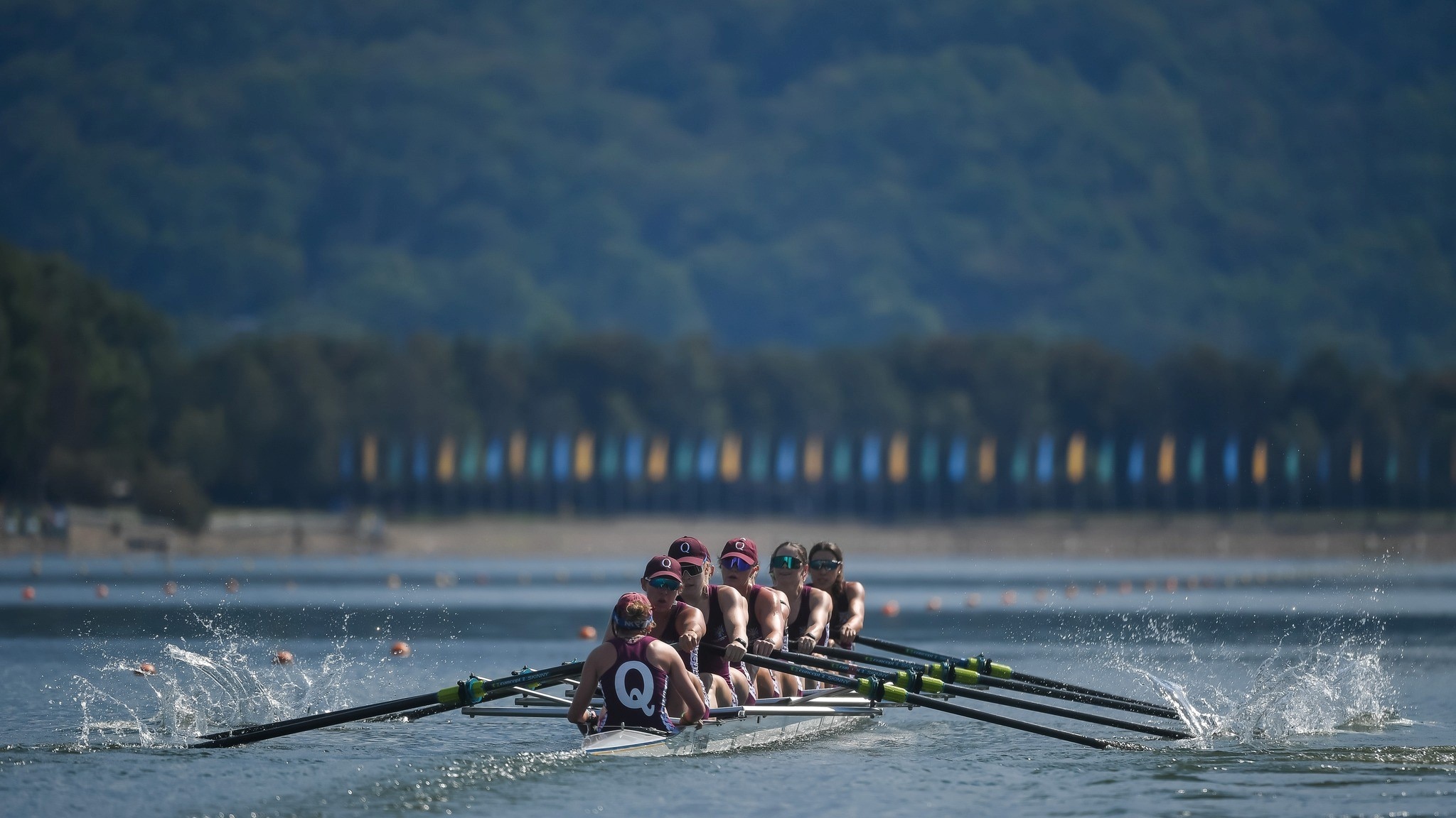 Uma foto ampla de uma equipe feminina de remo durante uma corrida. Eles estão vestindo marrom e a cox tem um 'Q' nas costas