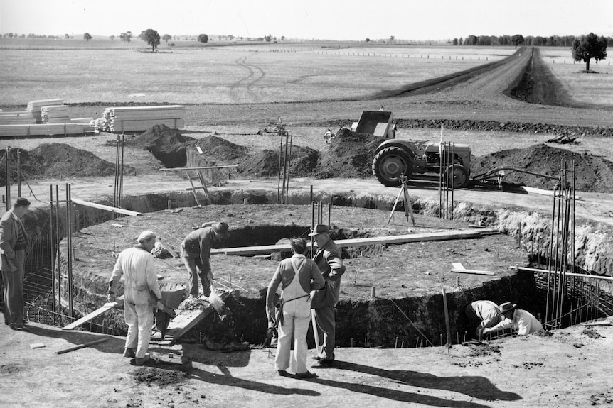 A black and white photo showing a group of people standing in front of a hole in the ground.