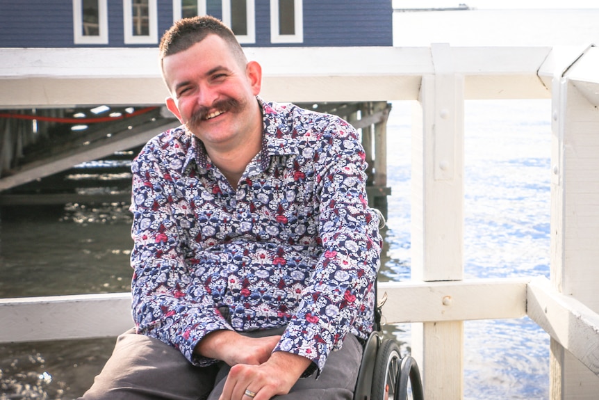 A smiling man with a large moustache, in a wheelchair, on a jetty.