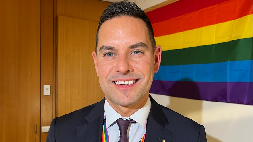 a man looking and smiling at the camera with a rainbow flag in the background