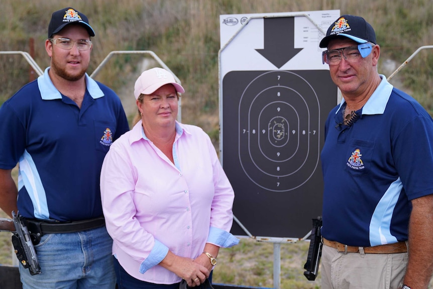 A family of competitive shooters stand next to a target. Two men are wearing protective glasses, with guns slung on their hips