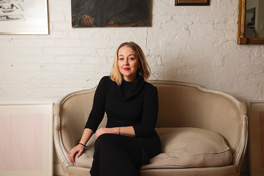 A woman wearing a black outfit sitting on a cream sofa wearing red lipstick.