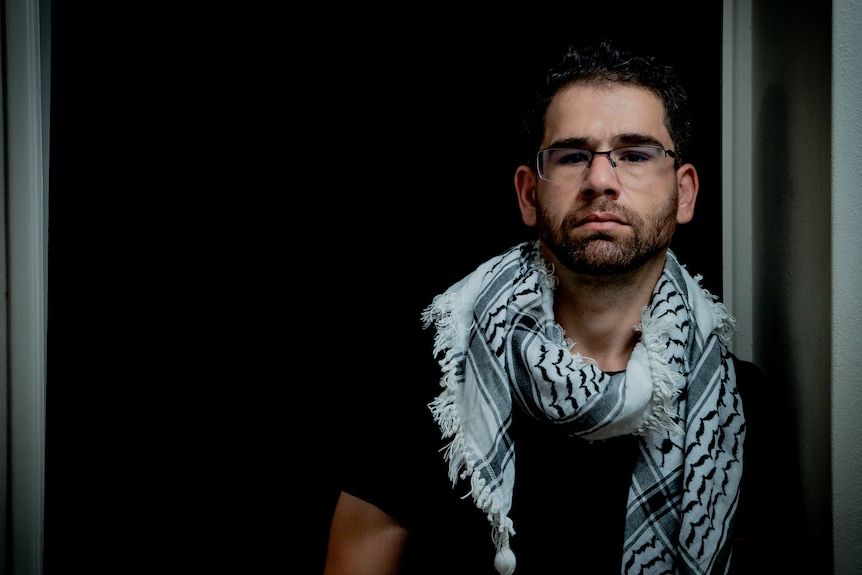 Ahmed is leaning against a wall, wearing a black shirt with a white scarf