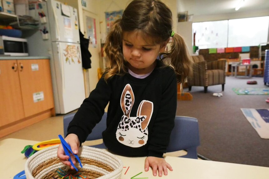 A little girl with pigtails concentrates hard as she plays with tweezers and a basket of coloured match sticks.