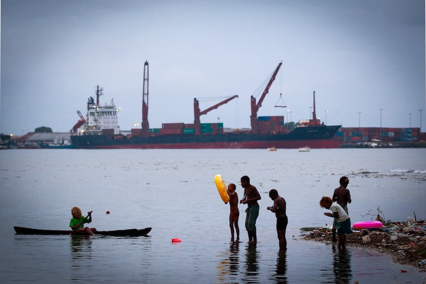 A group of children play at the water's edge with tankers in the background 