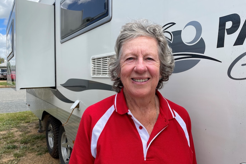A smiling older woman wearing a bright-coloured shirt stands in front of a caravan.