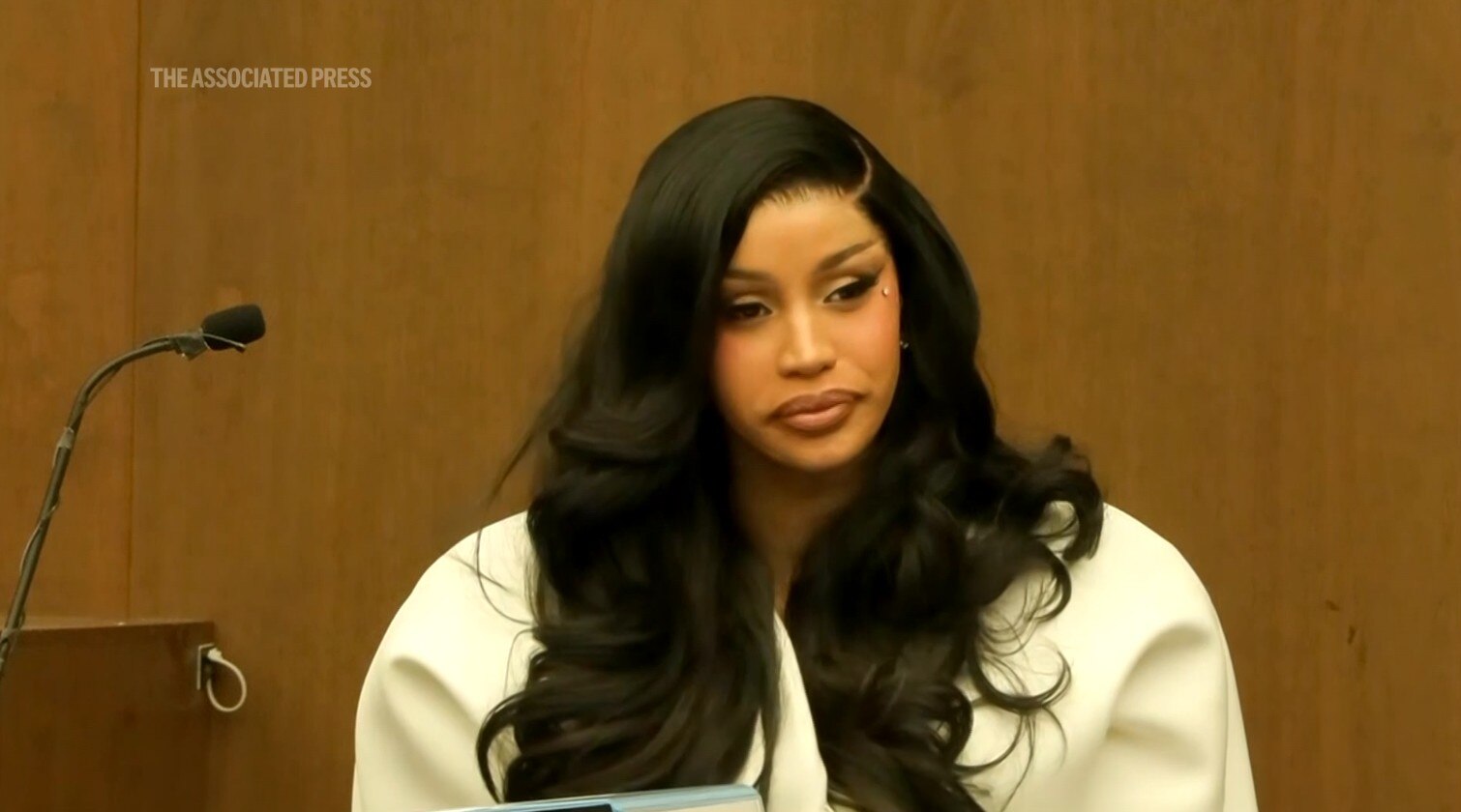 Cardi B sits in a witness box with long dark curly hair with a neutral facial expression. 