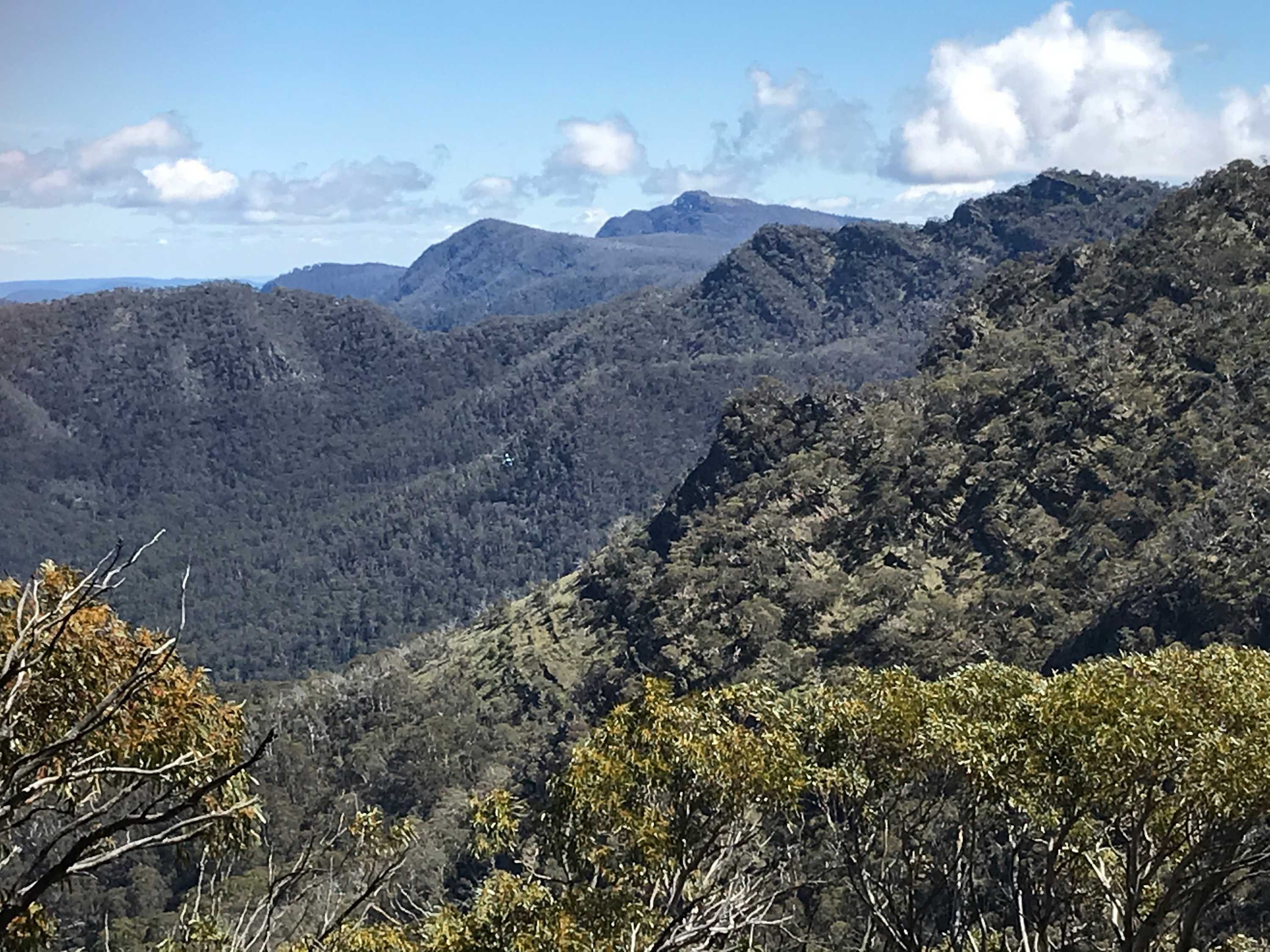 Green mountains take up the picture, a tiny helicopter can just be seen flying over a valley in the middle of the picture.