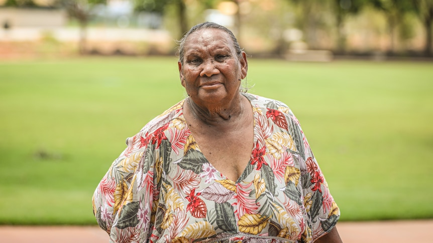 A female Indigenous elder looks at the camera with a stern expression, wears printed blouse, with green, red, yellow leaves.