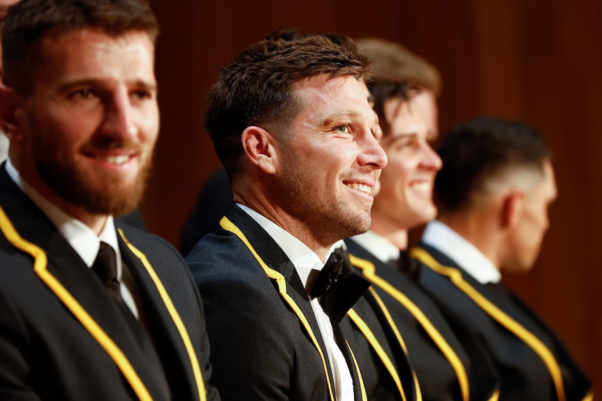 An AFL player wearing a blazer smiles as he stands on stage alongside teammates during a presentation.