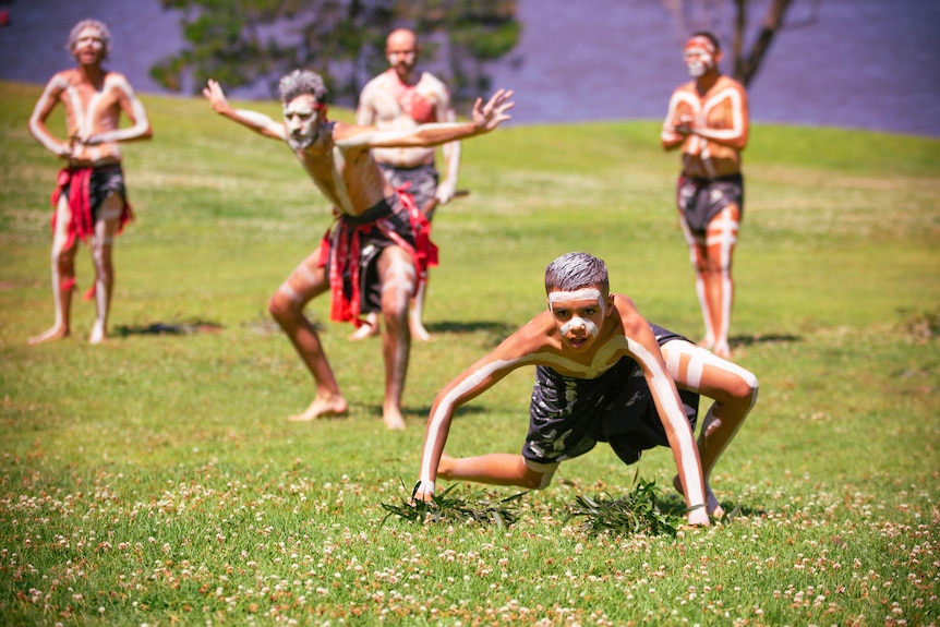 Five members of the Gadhungal Murring group wearing body paint and traditional dress, dancing on the grass next to a river.