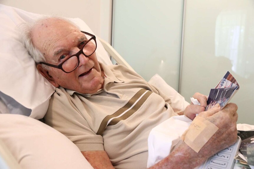 Elderly man with glasses and a bandage on his hand lies in a bed while holding photos.