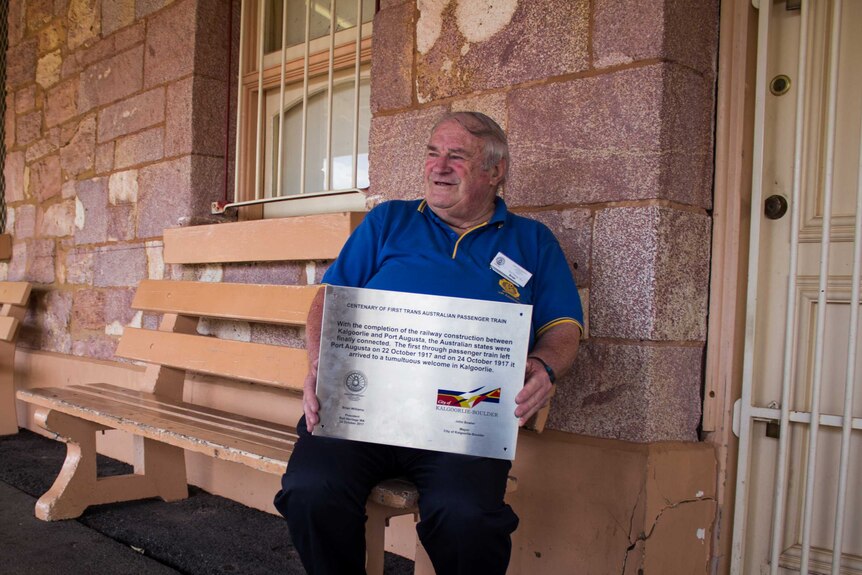 Brian Williams sits at Kalgoorlie Train Station, holding a commemorative plaque.
