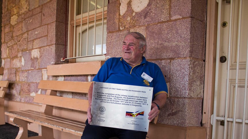 Brian Williams sits at Kalgoorlie Train Station, holding a commemorative plaque.